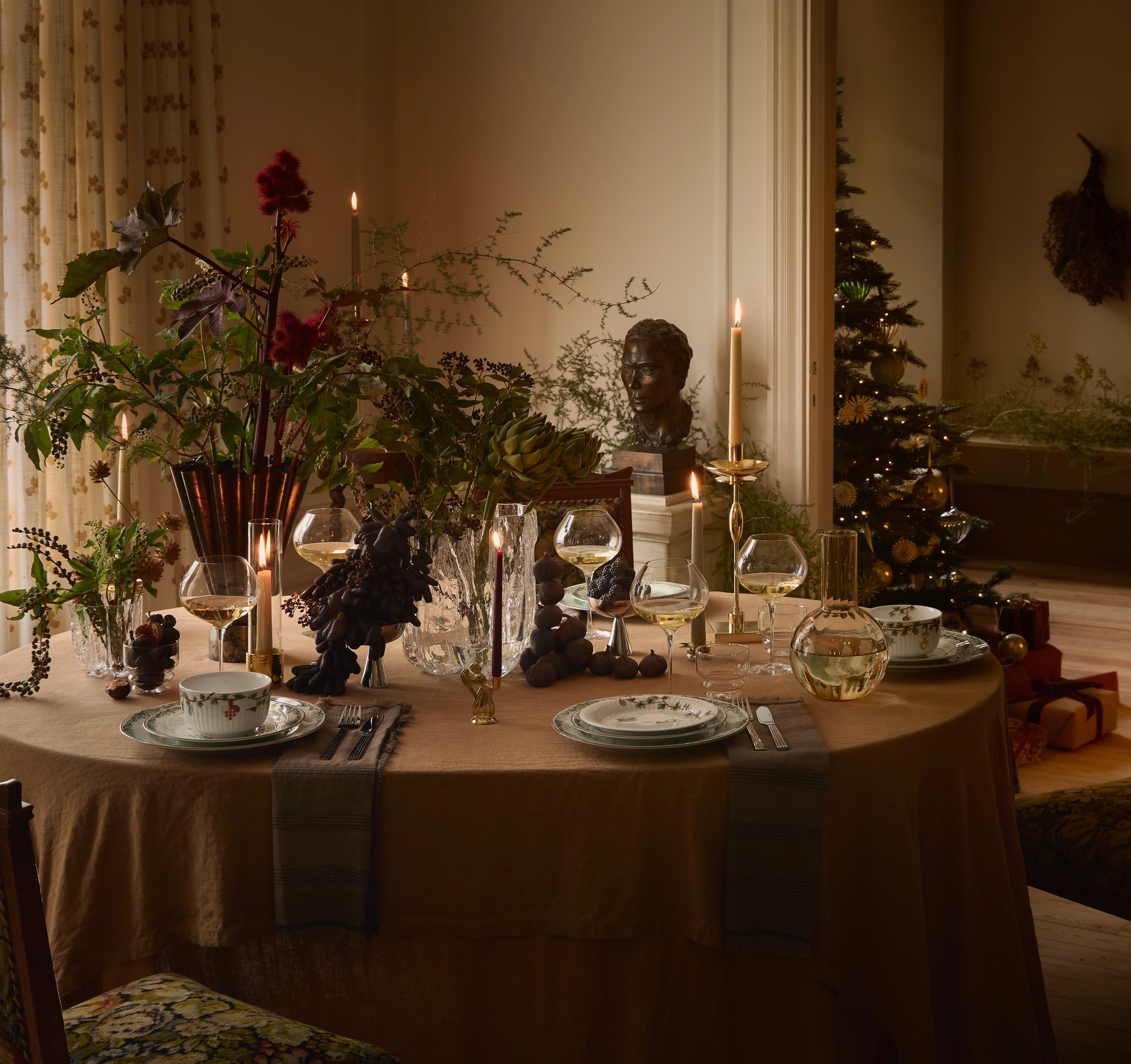Ett elegant dukat matbord med en dramatisk mittdekoration i lövverk och ljus. En tänd julgran syns genom en öppen dörröppning.