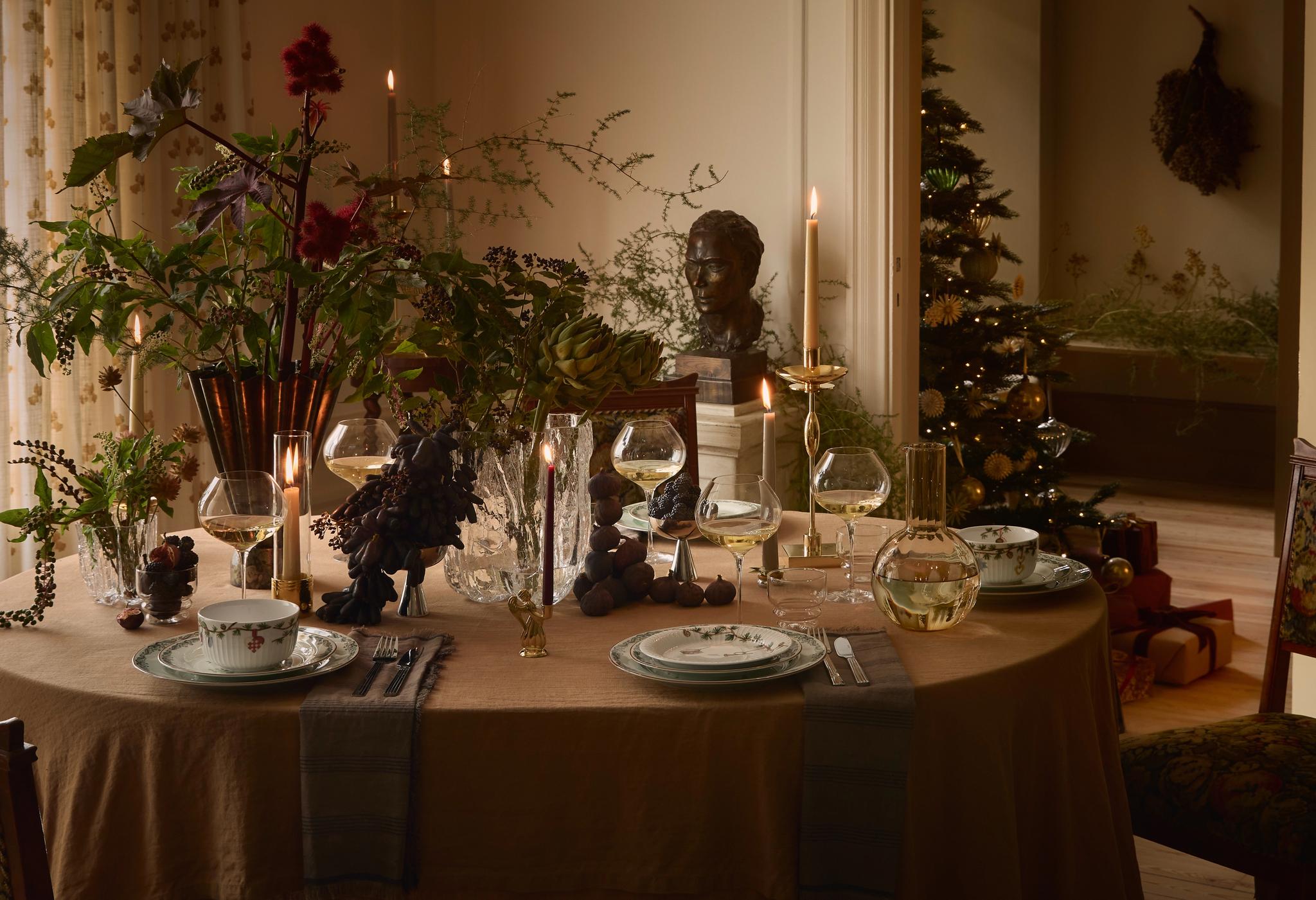 Ett elegant dukat matbord med en dramatisk mittdekoration i lövverk och ljus. En tänd julgran syns genom en öppen dörröppning.
