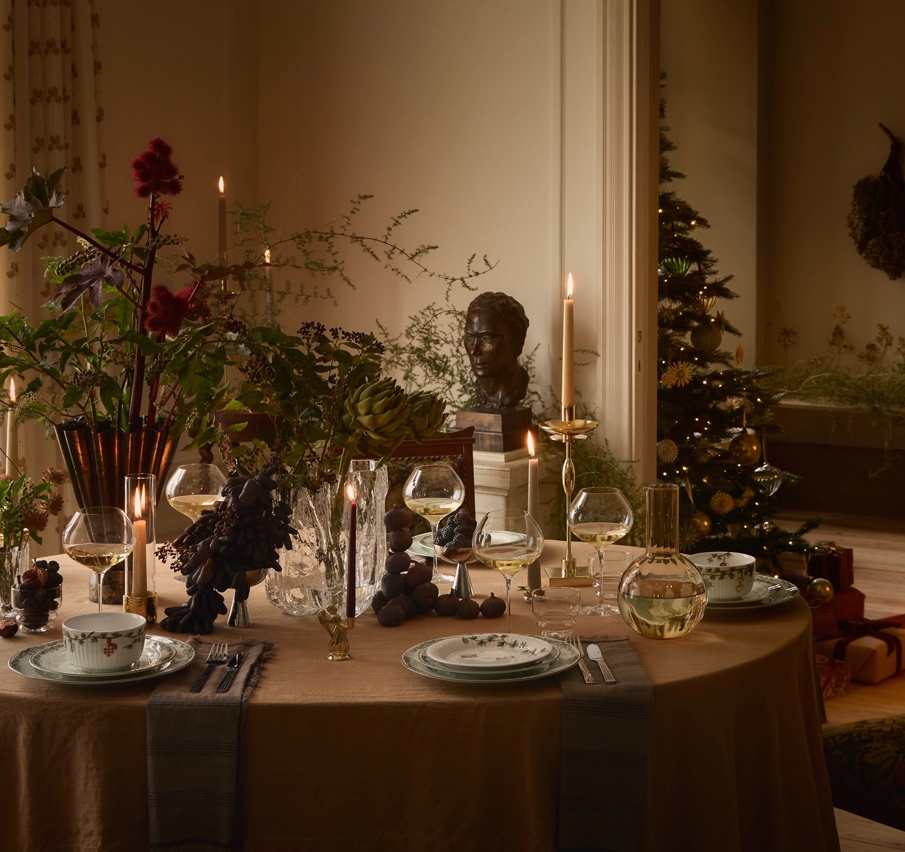 An elegantly set dining table with a rich floral centerpiece and lit candles, with a festive Christmas tree visible through an open doorway in the background.