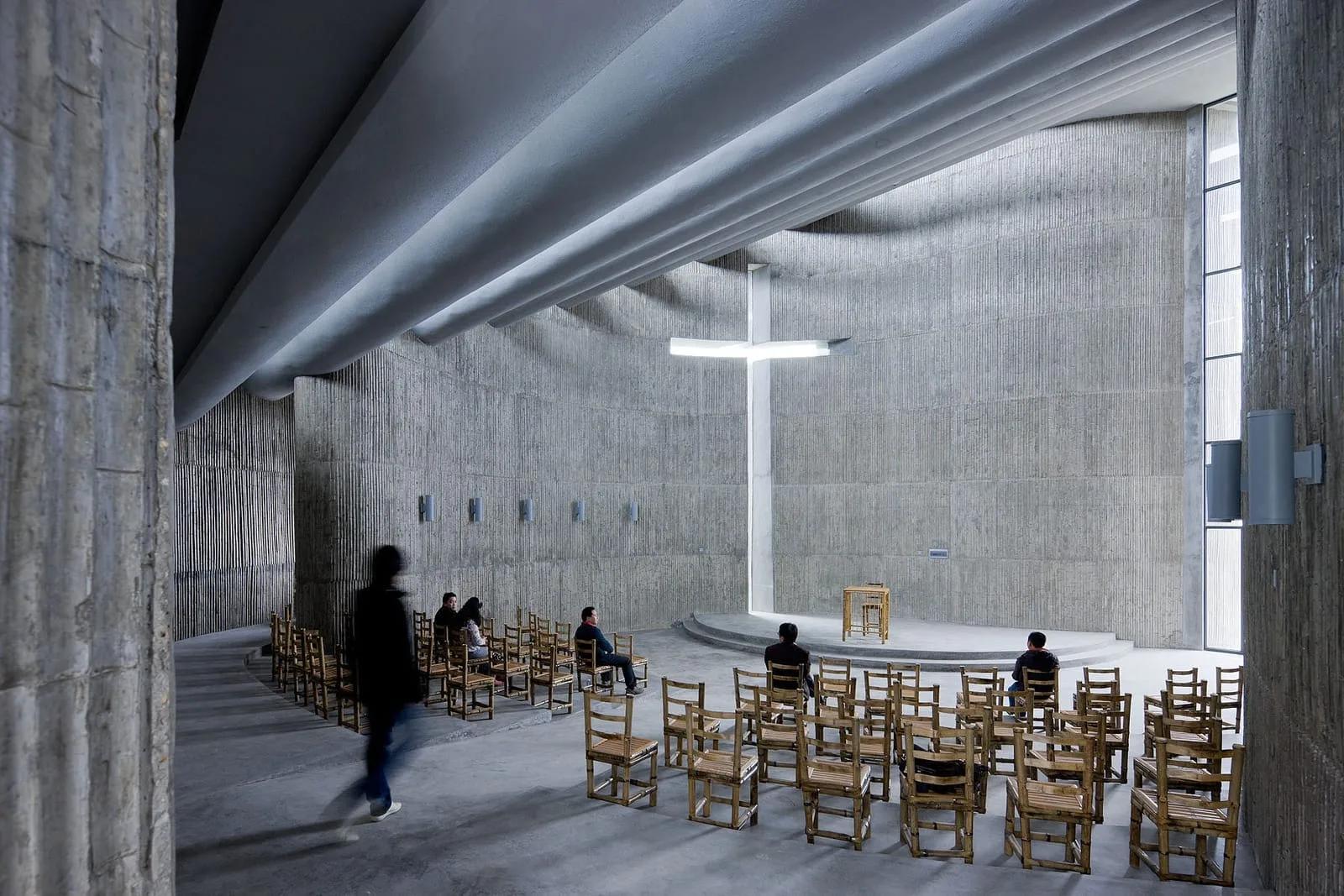 Interior de igreja em concreto com parede curva, cadeiras de madeira e uma cruz de luz recortada na parede do fundo
