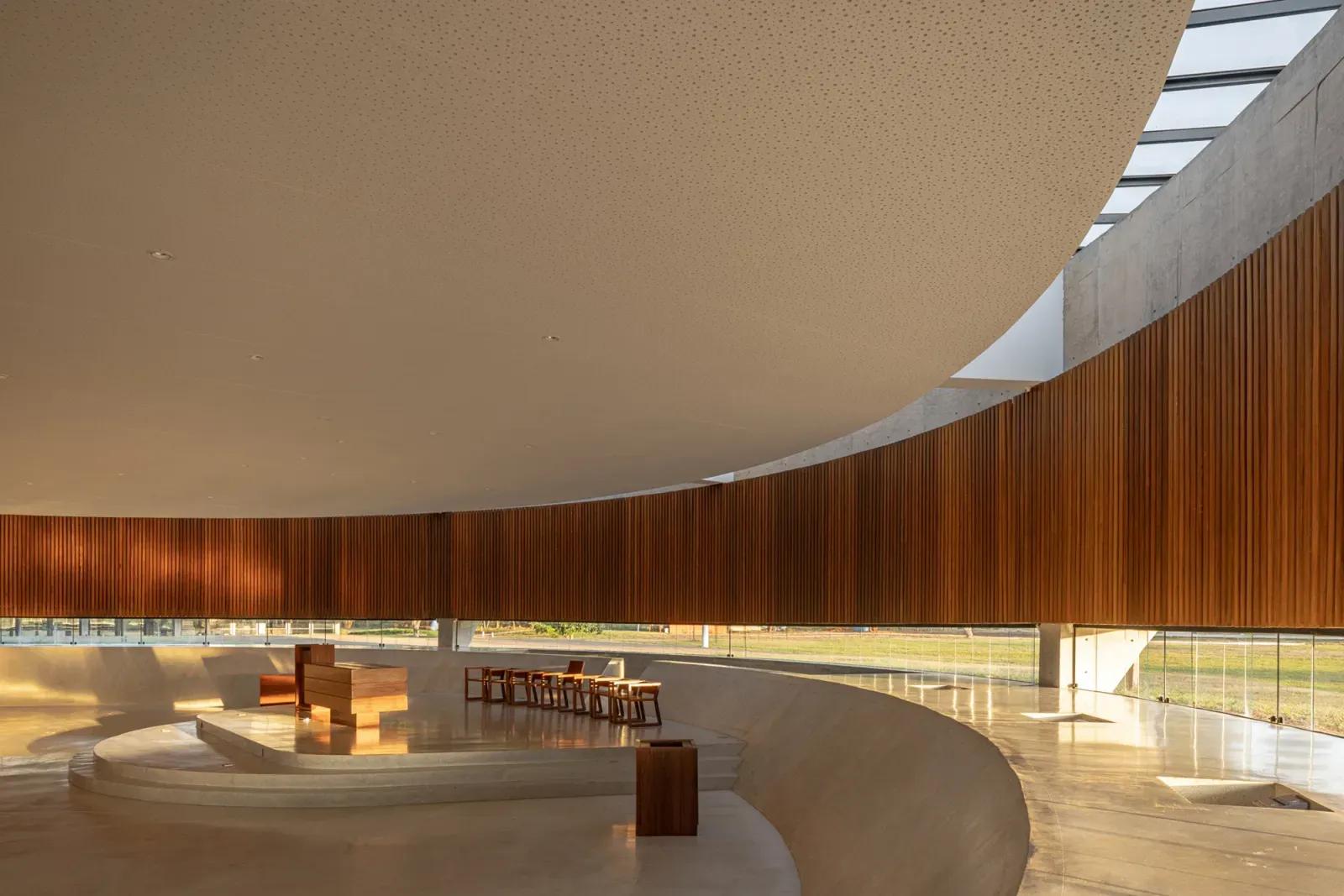Paróquia Sagrada Família, interior Interior circular com parede revestida de madeira, bancos baixos e luz entrando por um anel na cobertura