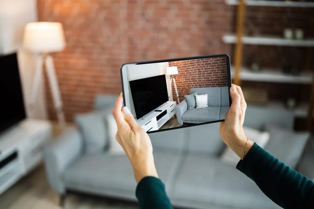 Vistoria com aplicativo Mãos seguram um tablet que mostra a imagem da sala de estar ao fundo, com sofá, estante e luminária