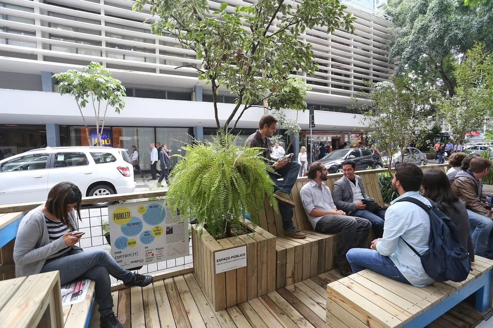 Parklet de madeira com bancos e vegetação em São Paulo Pessoas sentadas em bancos de madeira de um parklet com plantas, na calçada diante de um prédio, em São Paulo