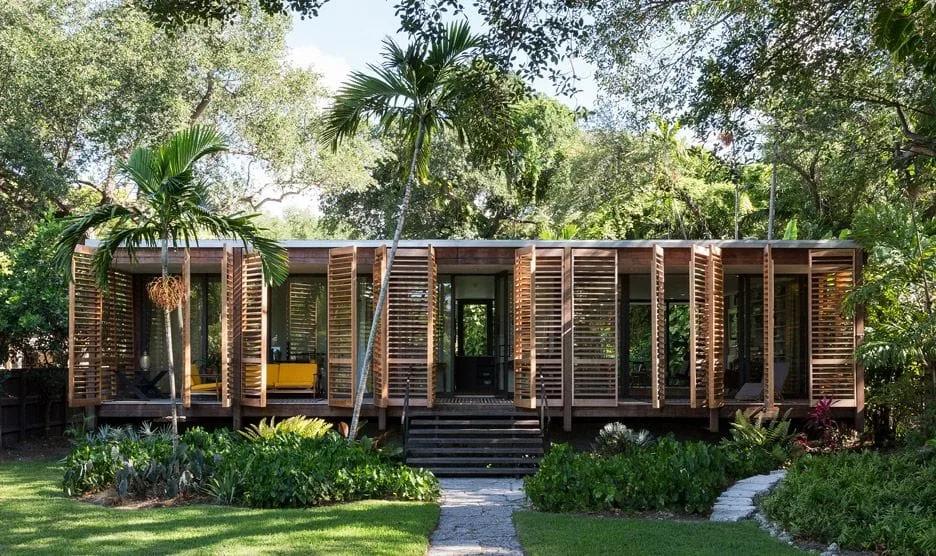 Casa térrea de madeira com painéis móveis na fachada, cercada por jardim tropical e árvores