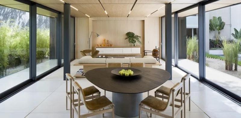 Sala integrada da SysHaus com mesa de jantar escura, cadeiras de madeira e grandes panos de vidro voltados para o jardim