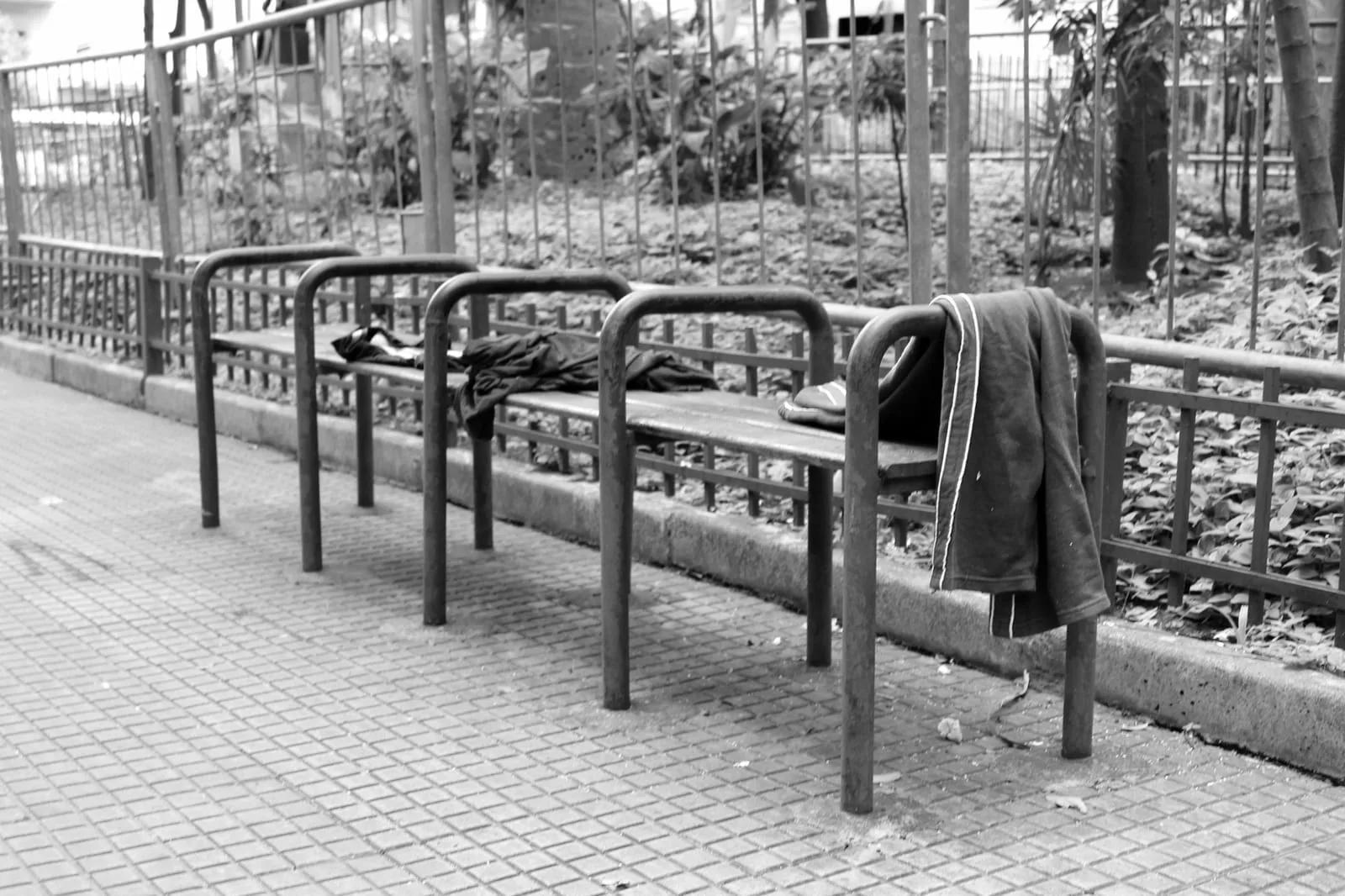 Banco público de metal dividido em assentos individuais por barras curvas, com roupas deixadas sobre ele, em foto preto e branco