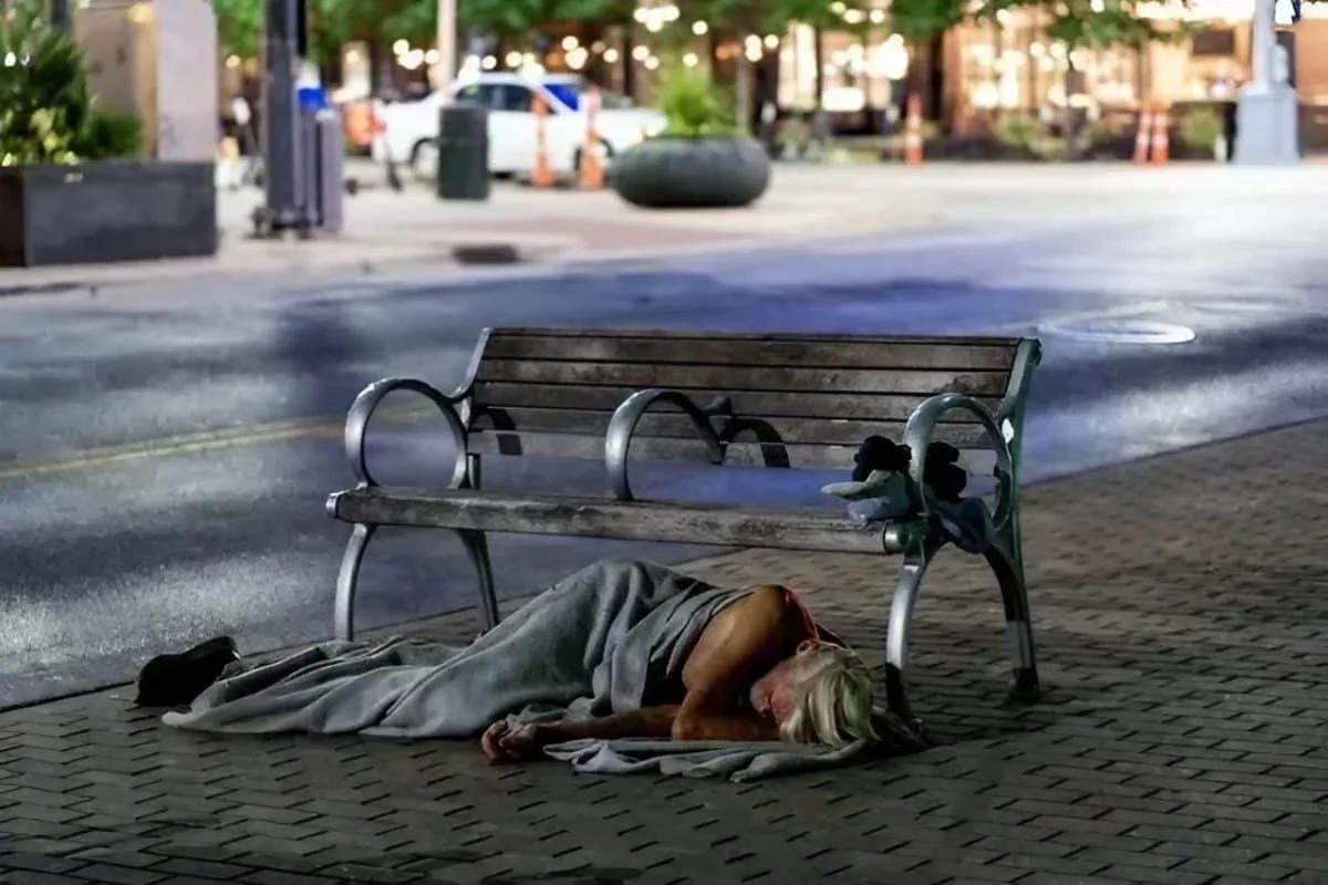 Pessoa em situação de rua dorme no chão ao lado de banco com divisórias Pessoa deitada na calçada, coberta por um cobertor, ao lado de um banco de praça com braços metálicos que dividem o assento