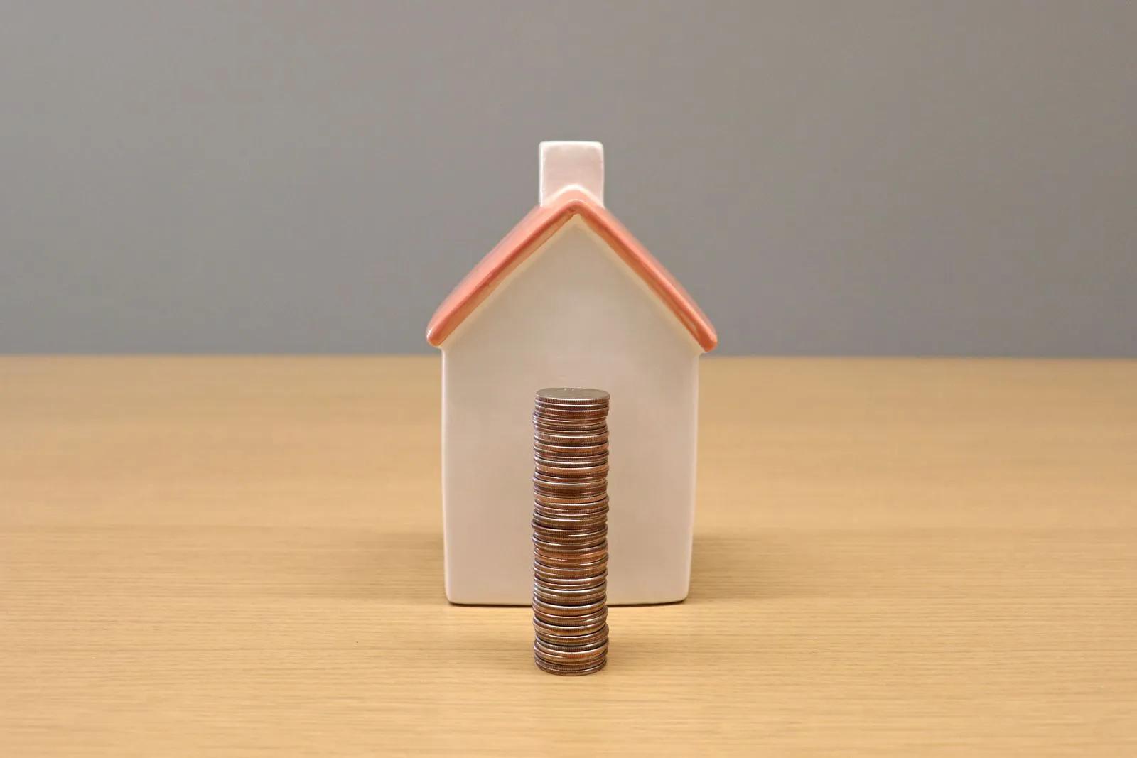 Casinha de porcelana com uma pilha de moedas à frente, sobre uma mesa de madeira