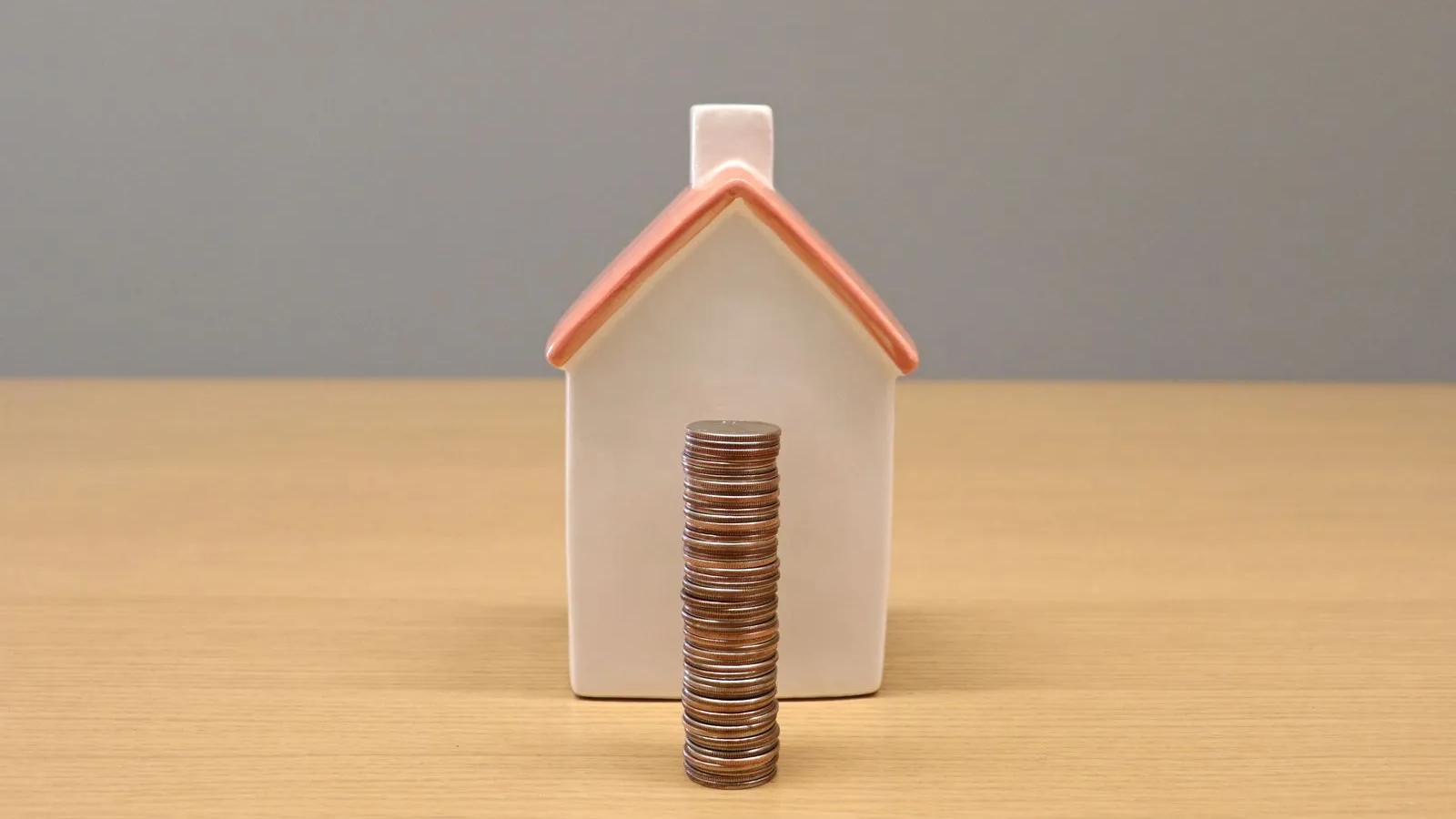 Casinha de porcelana com uma pilha de moedas à frente, sobre uma mesa de madeira