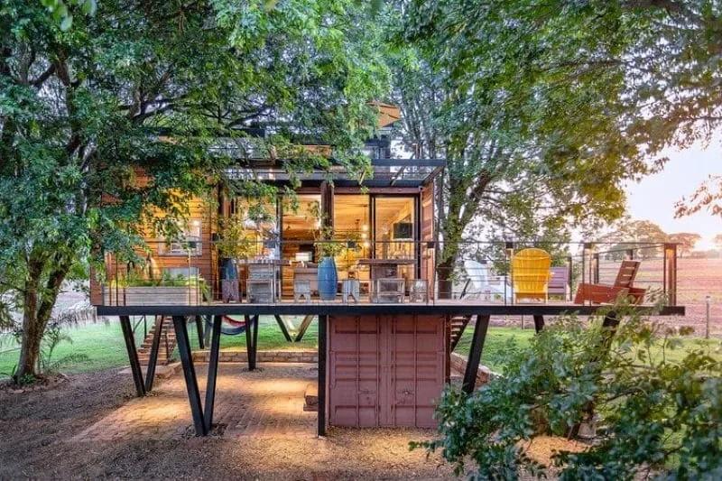 Casa container marrom elevada sobre pilares metálicos, com deque de madeira e árvores ao redor ao entardecer