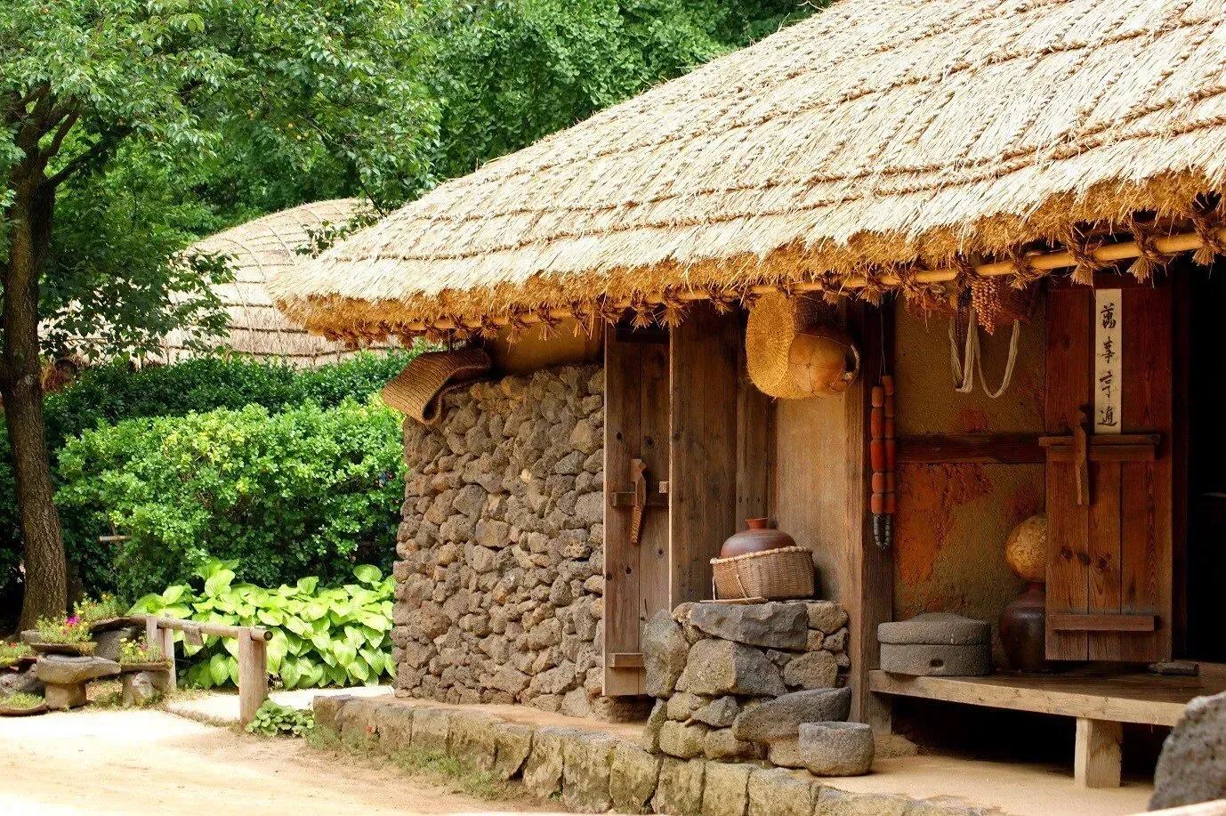 Casa tradicional coreana com telhado de palha, parede de pedras e estrutura de madeira, entre árvores