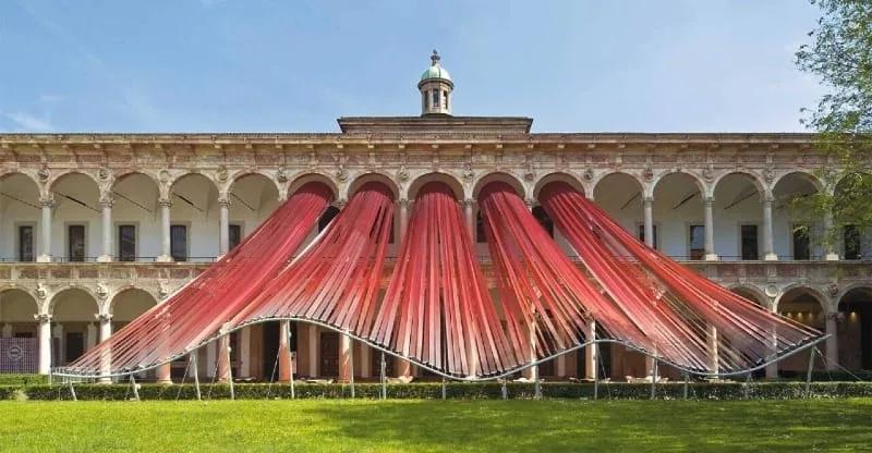 Instalação de arcos vermelhos translúcidos montada no pátio de um palácio histórico de Milão durante o Fuorisalone