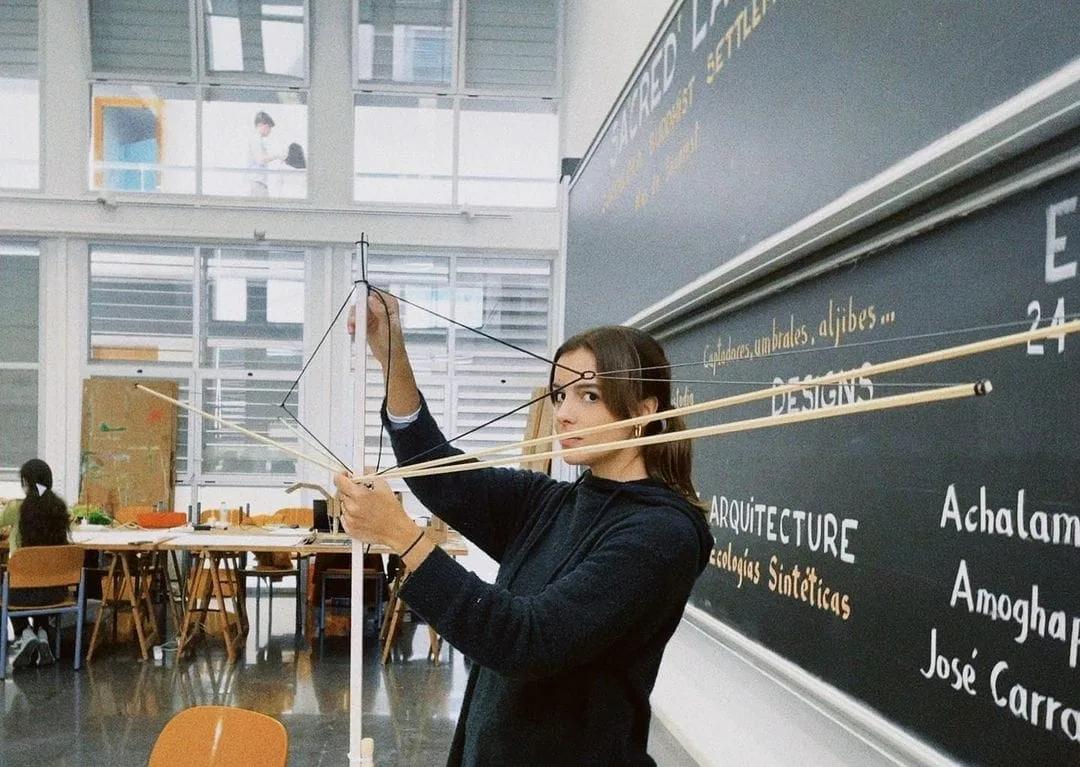 Estudante segura uma estrutura de varetas de madeira e fios diante de um quadro-negro em um ateliê de arquitetura