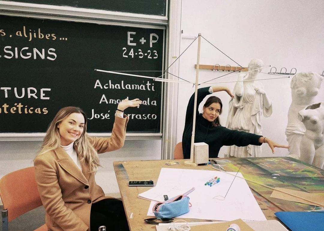 Duas estudantes sorriem e apontam para uma estrutura de varetas sobre a mesa de um ateliê com quadro-negro e esculturas