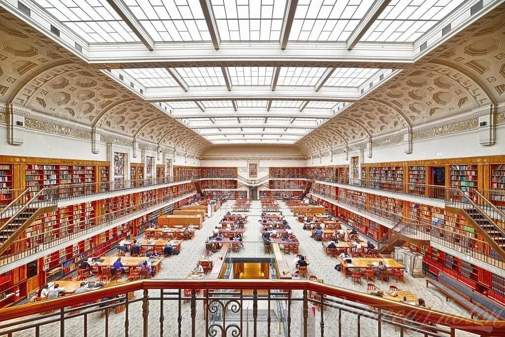 Sala de leitura Sala de leitura com teto de vidro, mesas longas e estantes em galerias nas paredes