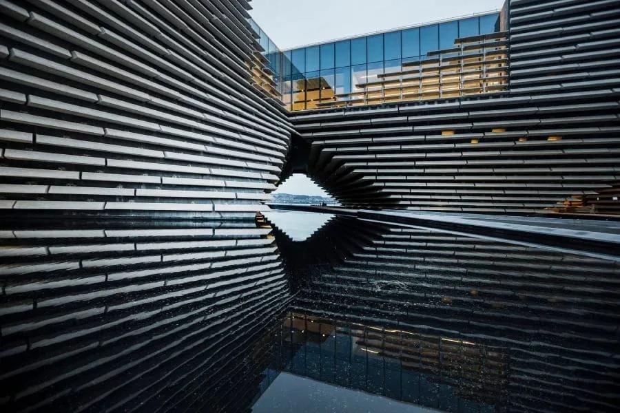Fachada de lâminas de concreto do V&A Dundee com a passagem central refletida na água