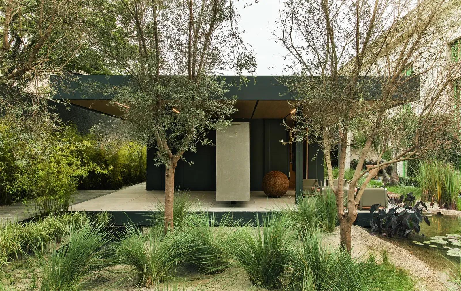 Fachada escura da SysHaus vista do jardim, com oliveiras, touceiras de capim, bambus ao fundo e lago com plantas aquáticas