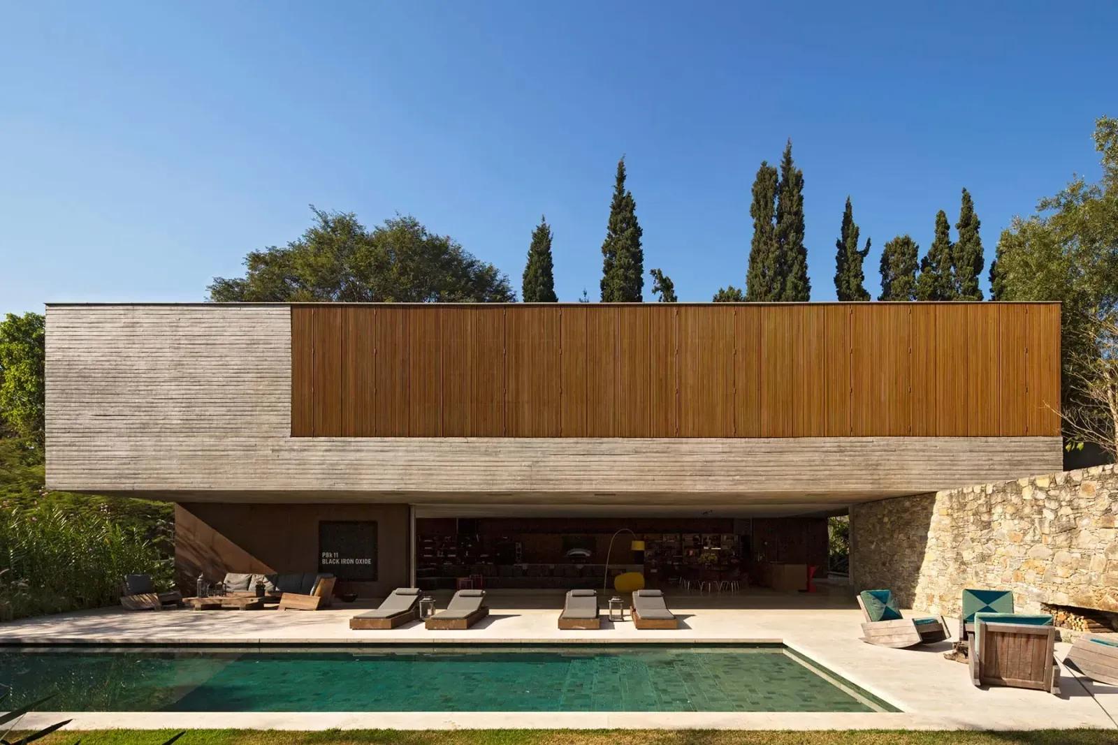 Casa com volume superior revestido de ripas de madeira sobre térreo de vidro, piscina e muro de pedra