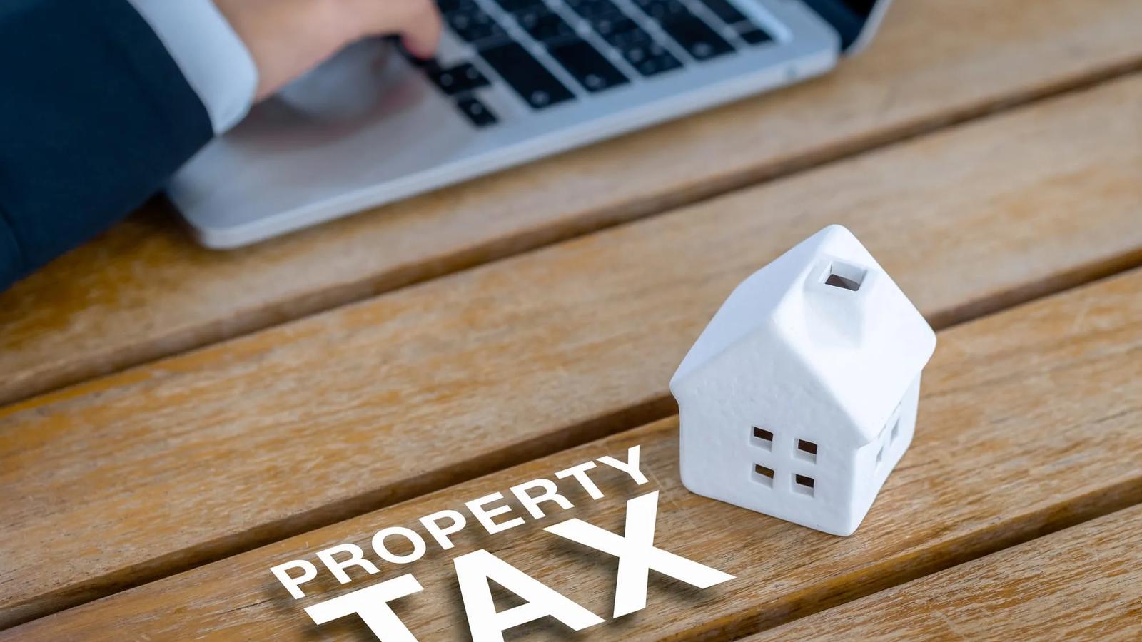 Casa em miniatura sobre mesa de madeira com as palavras property tax e uma pessoa usando notebook ao fundo