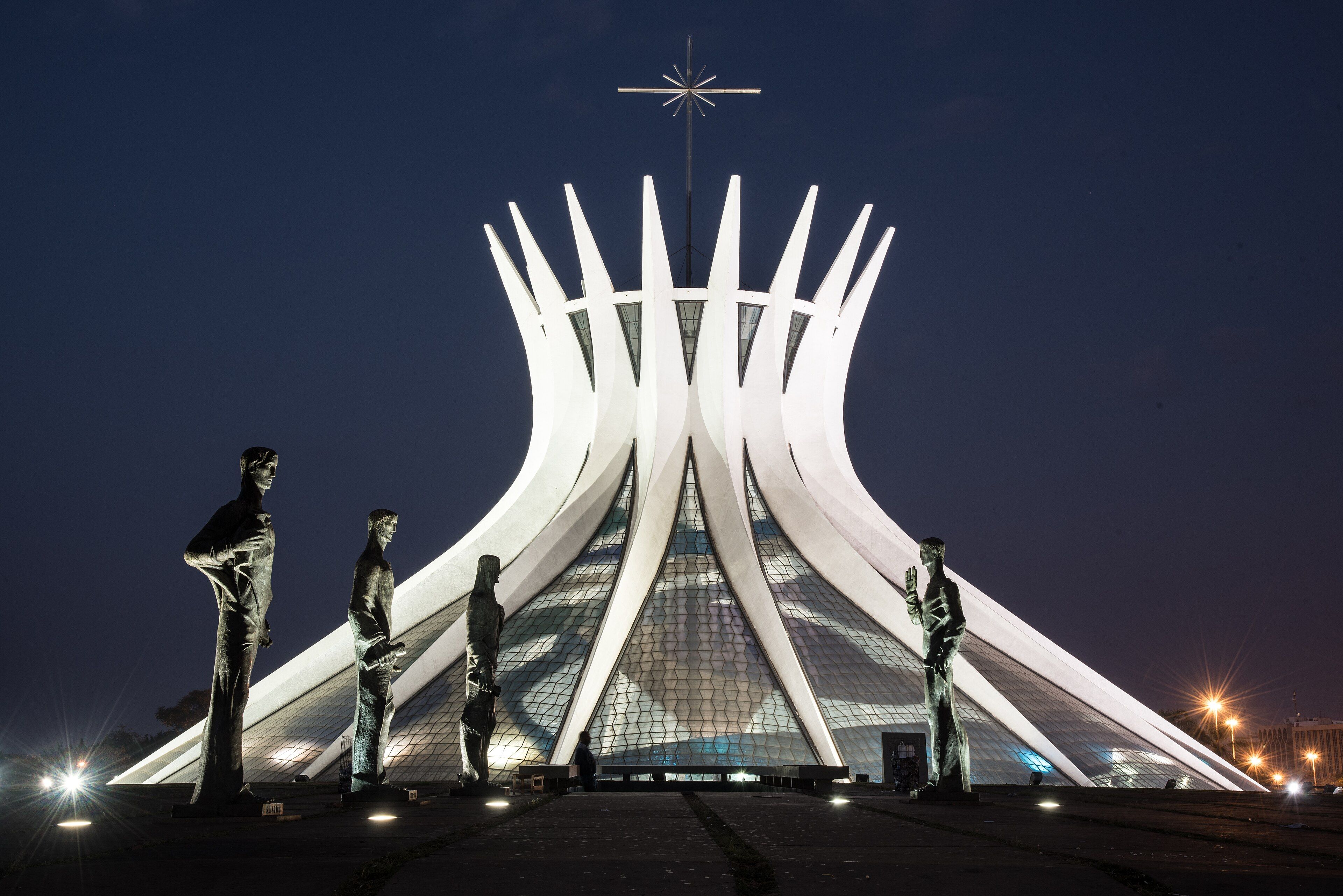 Catedral de Brasília iluminada à noite, com as colunas curvas de concreto e quatro estátuas de bronze dos evangelistas na praça em primeiro plano