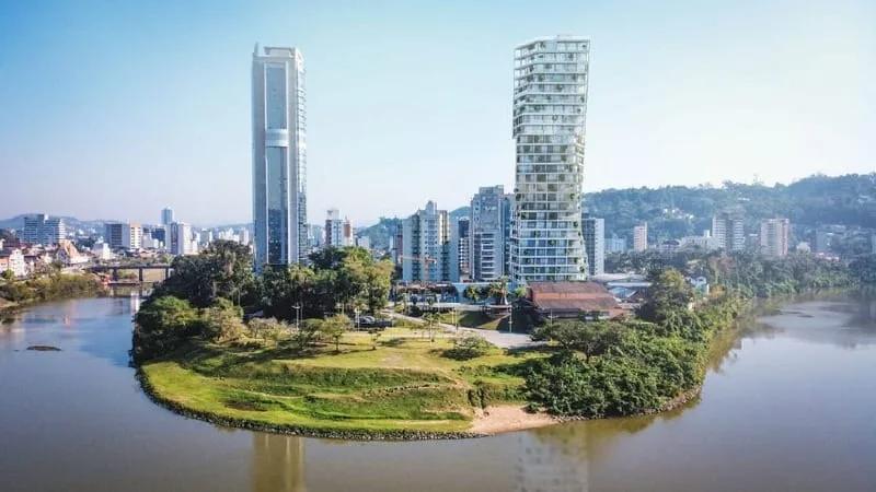 Projeção de edifícios altos em Blumenau Montagem com dois edifícios altos às margens do rio Itajaí-Açu, em Blumenau, com um gramado em primeiro plano e a cidade ao fundo