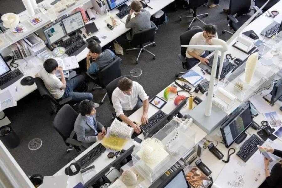 Escritório de arquitetura com várias pessoas trabalhando em computadores e mesas cheias de papéis e maquetes