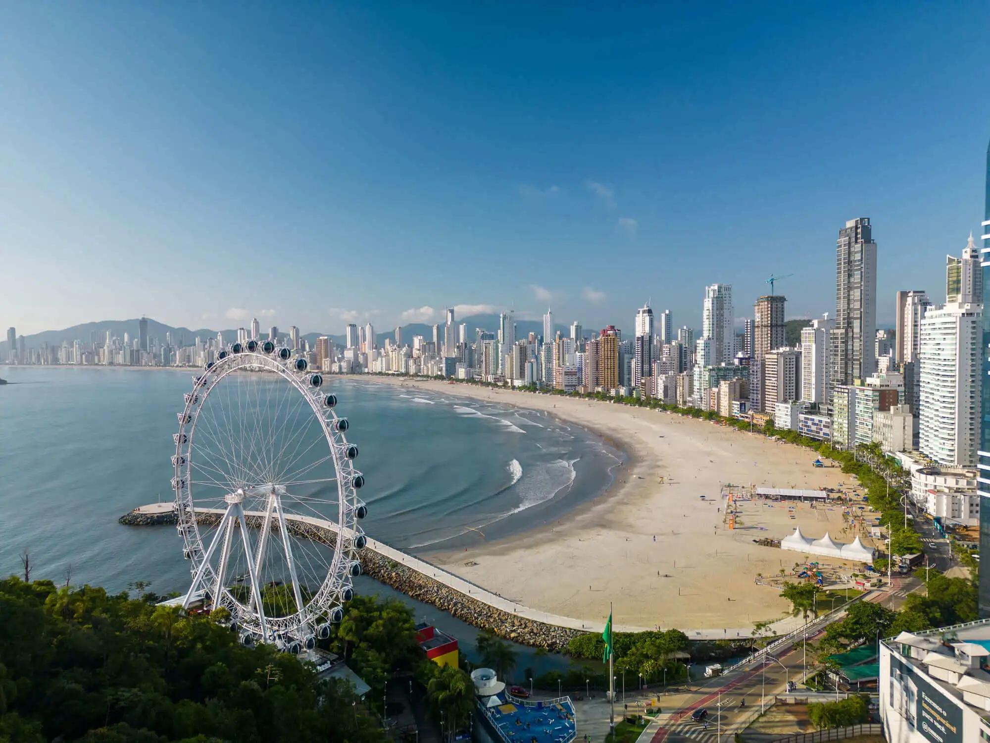 Foto da orla de Balneário Camboriú, aparecendo predios, praia e roda gigante da cidade.