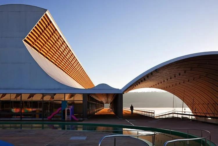 Coberturas curvas com forro de madeira sobre área de piscina e parque infantil, à beira do lago