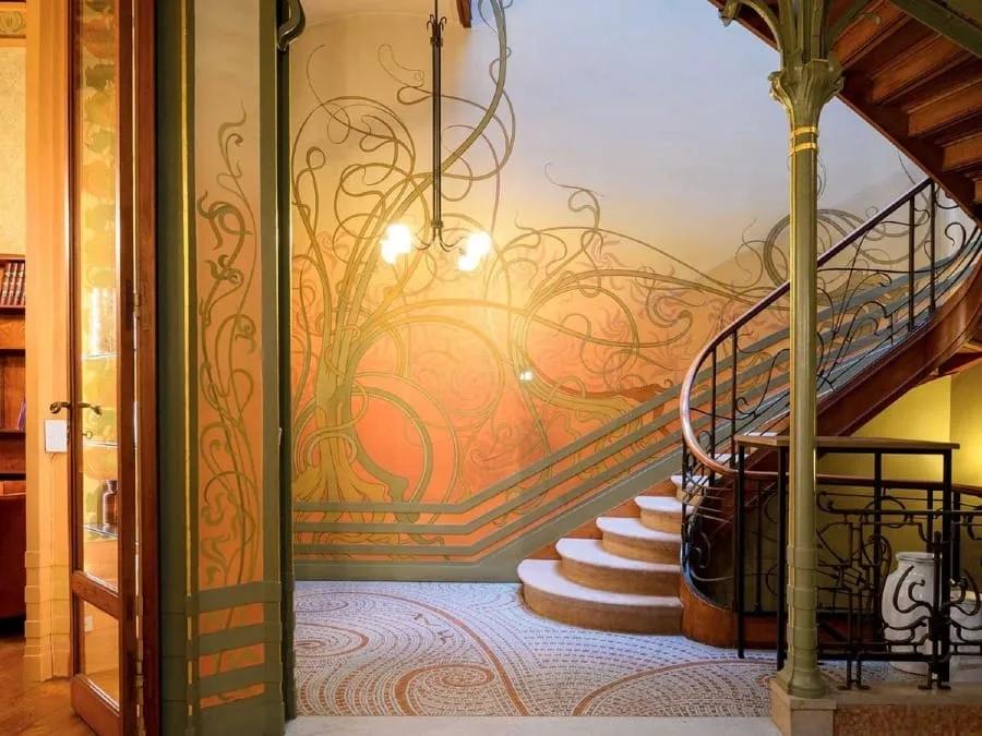 Hall com parede pintada de linhas curvas, escada de madeira e coluna de ferro com ramificações