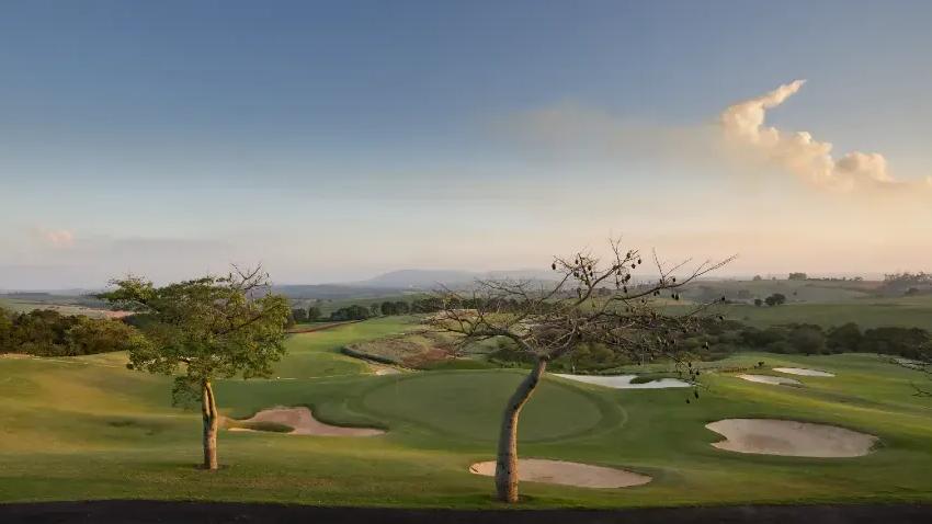 Campo de golfe ao entardecer Campo de golfe com greens, bancas de areia e duas árvores em primeiro plano, com colinas ao fundo ao entardecer