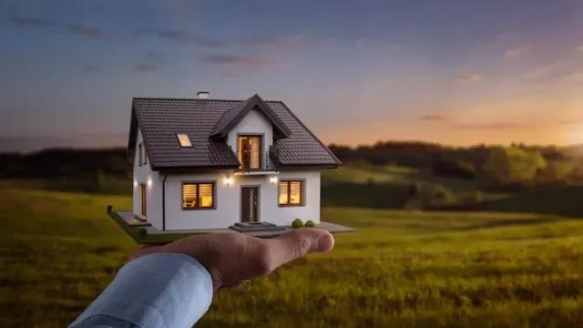 Mão segurando modelo de uma casa com um terreno rural ao fundo.