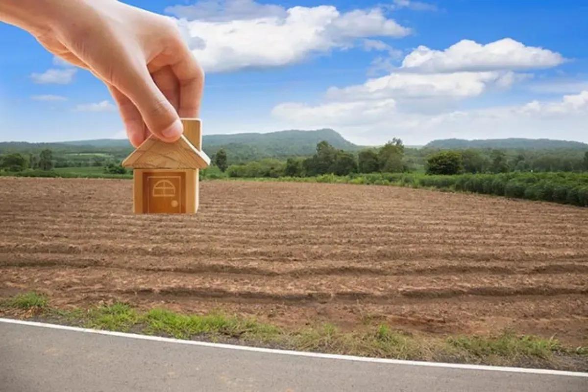 Mão segura uma pequena casa de madeira sobre um terreno de terra arada, com vegetação e morros ao fundo