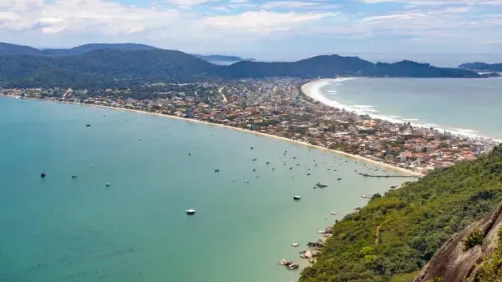 Vista aérea de uma enseada de água esverdeada com barcos ancorados, praia em curva e casario entre morros cobertos de mata.