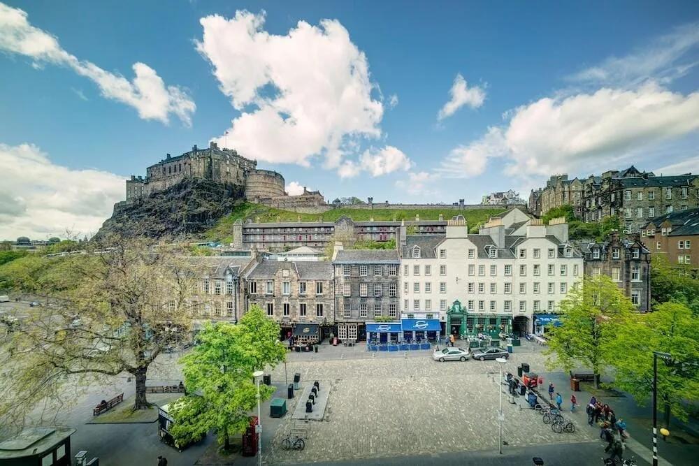 Praça do Grassmarket com prédios de pedra e o castelo sobre a rocha ao fundo, sob céu azul com nuvens