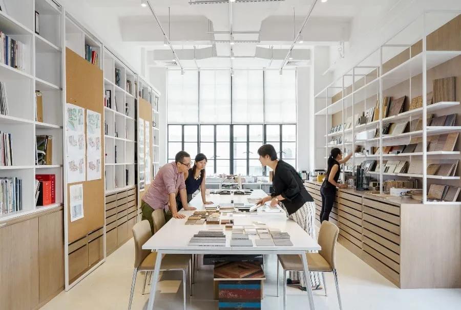 Estudantes e professores reunidos em torno de uma mesa com pranchas e cadernos, em sala cercada de estantes com livros