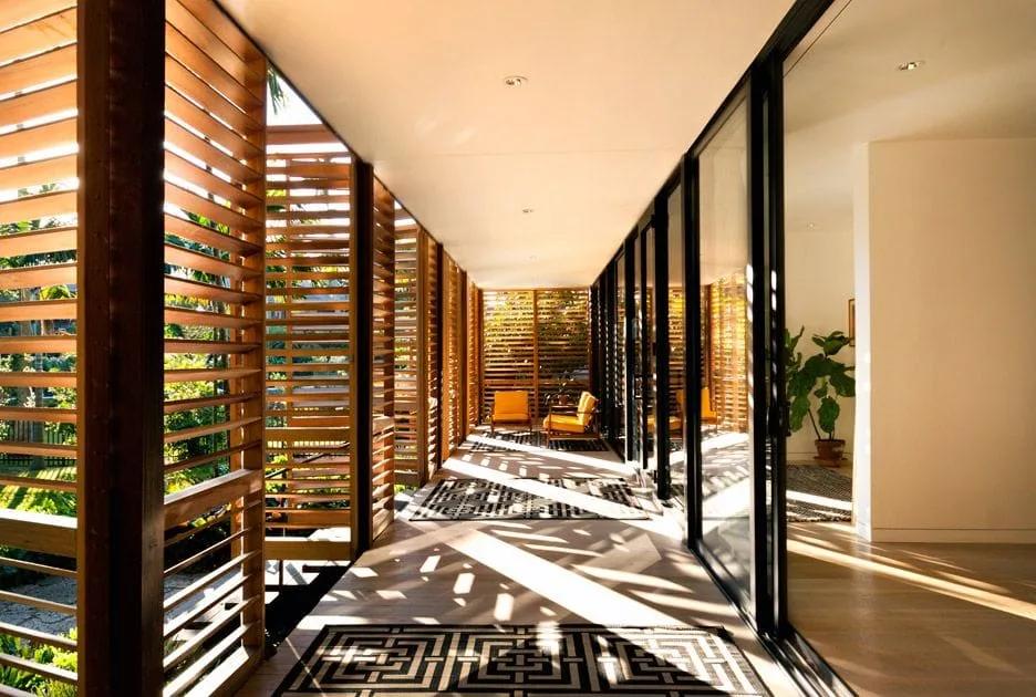 Varanda longa com painéis de madeira móveis projetando sombras no piso, entre a casa e o jardim tropical