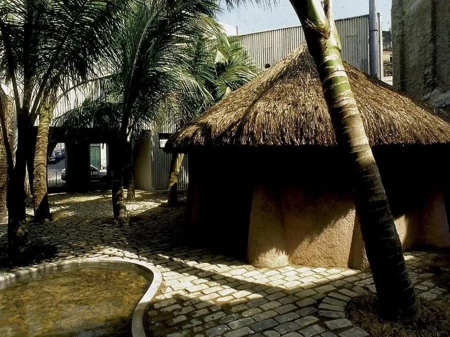 Pátio da Casa do Benin com cabana de palha, coqueiros e piso de pedra