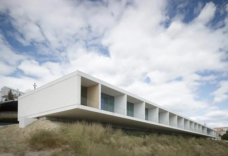 Edifício branco longo e horizontal com aberturas envidraçadas em sequência, sobre terreno com vegetação