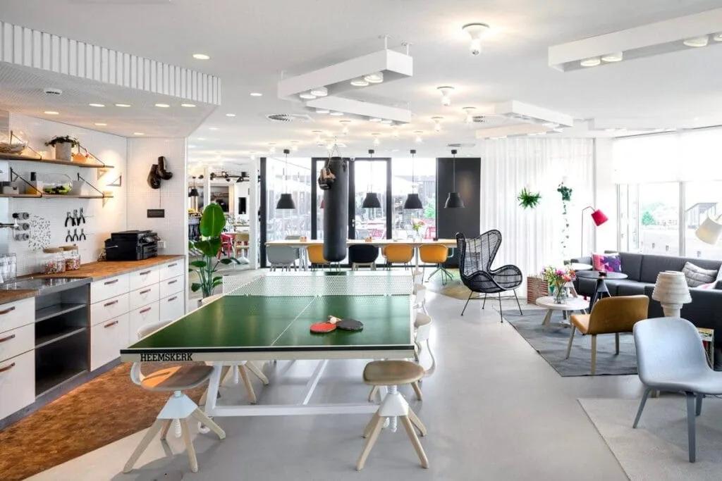 Área comum do Zoku, em Amsterdã Área comum de um coliving com mesa de pingue-pongue, cozinha, poltronas e plantas em um salão claro