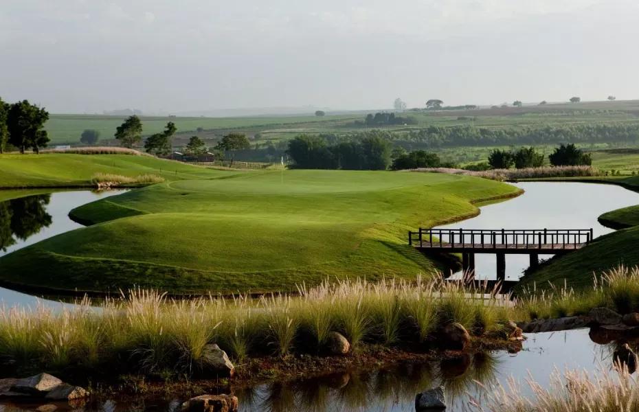 Campo de golfe com green cercado por lagos, ponte de madeira e gramíneas altas em primeiro plano, ao entardecer