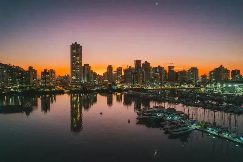 Pôr do sol sobre a Marina Itajaí, com barcos atracados e prédios iluminados refletidos no rio