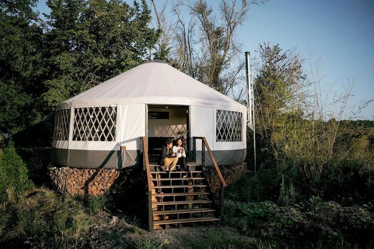 Yurta de cobertura clara com janelas de treliça, sobre um deck de madeira entre árvores