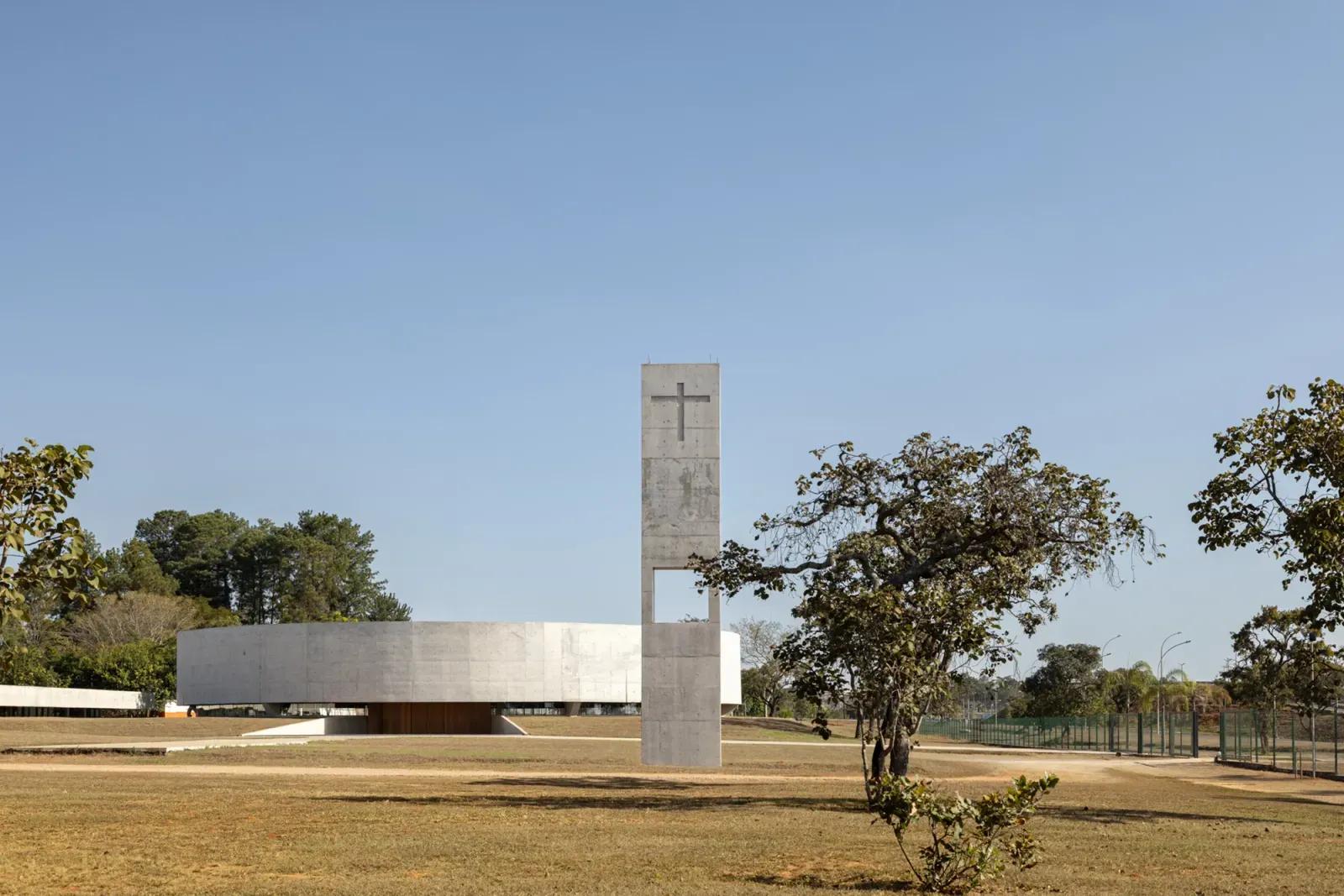 Torre sineira de concreto branco com uma cruz no alto e, ao fundo, a nave circular baixa num gramado amplo