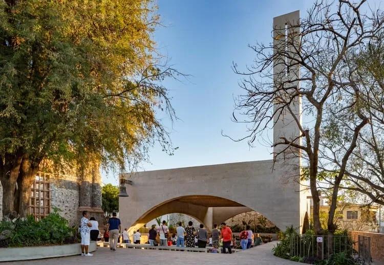 Grande arco de concreto aberto para uma praça com pessoas e árvores, ao lado de uma torre alta de concreto