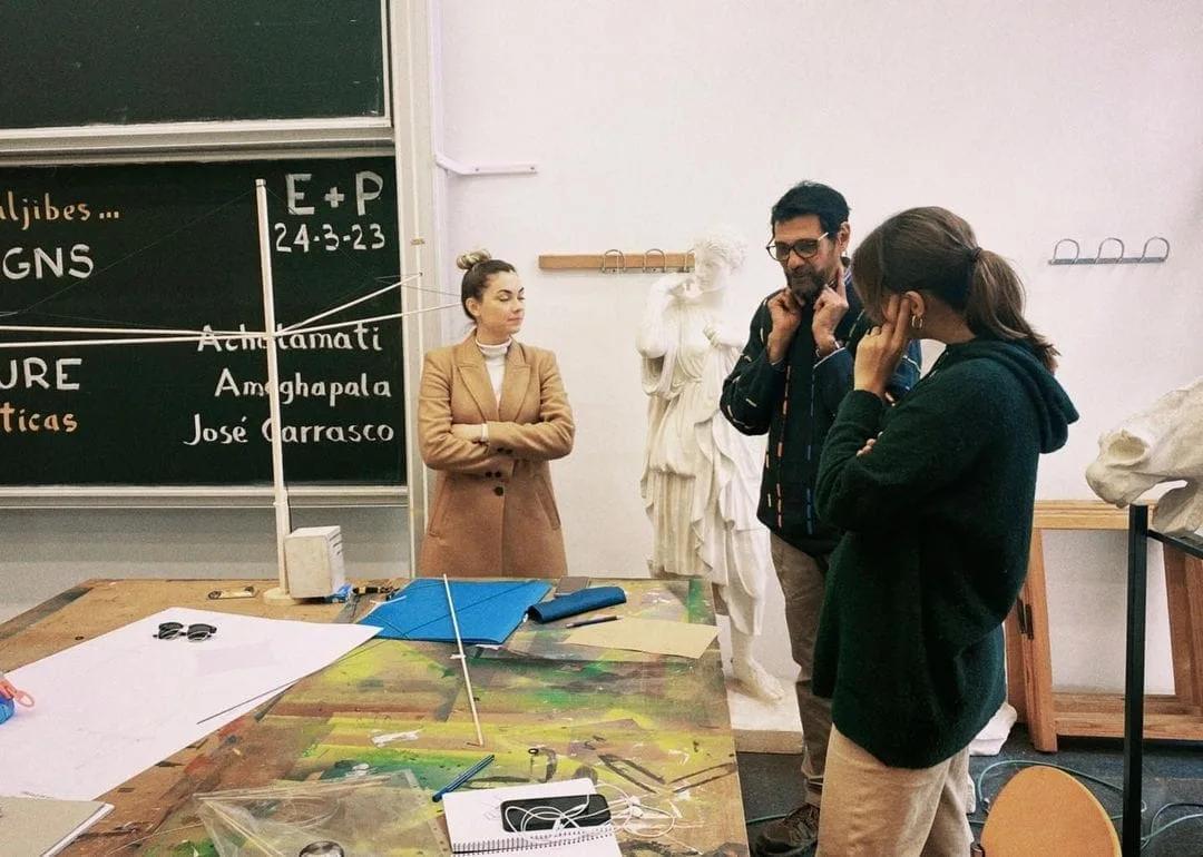 Três pessoas conversam em volta de uma mesa de ateliê com desenhos, diante de um quadro-negro e de esculturas de gesso