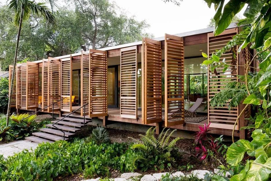 Brillhart House: casa elevada com painéis de madeira Casa térrea elevada do solo, com fachada de painéis de madeira abertos e jardim tropical à frente