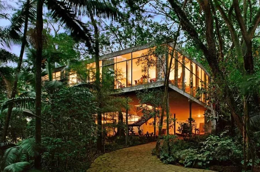 Casa de Vidro iluminada ao entardecer, cercada pela mata, com a escada de acesso sob a caixa envidraçada