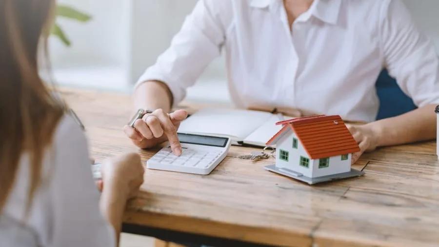 Duas pessoas à mesa de madeira com calculadora, papéis e uma pequena maquete de casa, fazendo contas de aluguel