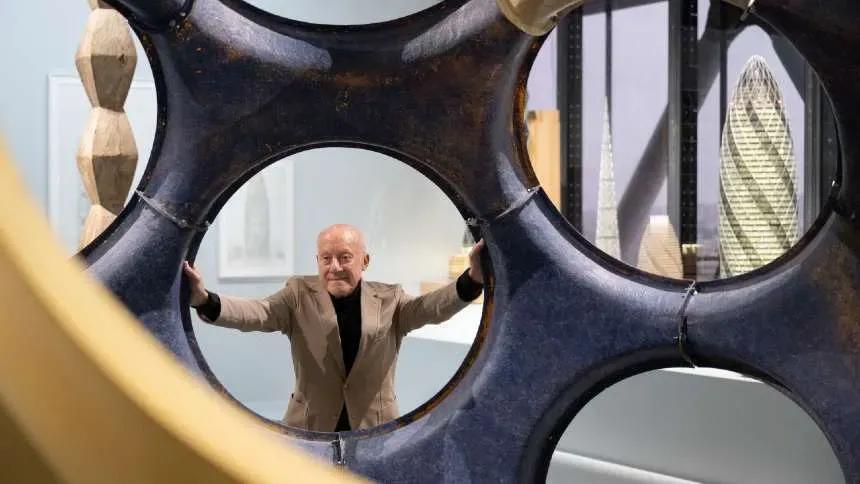 Norman Foster de braços abertos, visto através de uma escultura vazada, com maquetes de torres ao fundo, entre elas a do 30 St Mary Axe
