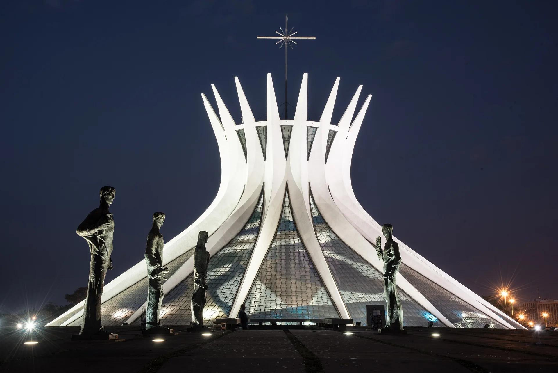 Catedral de Brasília iluminada à noite, com as colunas curvas de concreto e quatro estátuas de bronze dos evangelistas na praça em primeiro plano